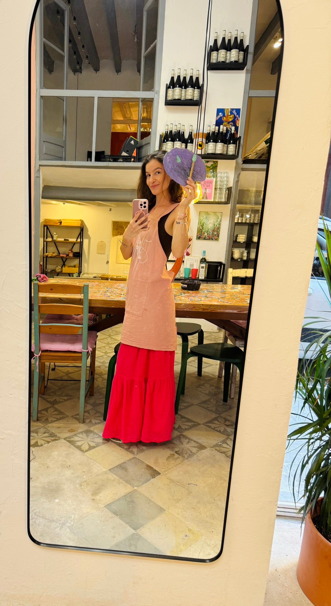 Person taking a mirror selfie wearing a pink anpron and red skirt in a room with shelves and furniture.
