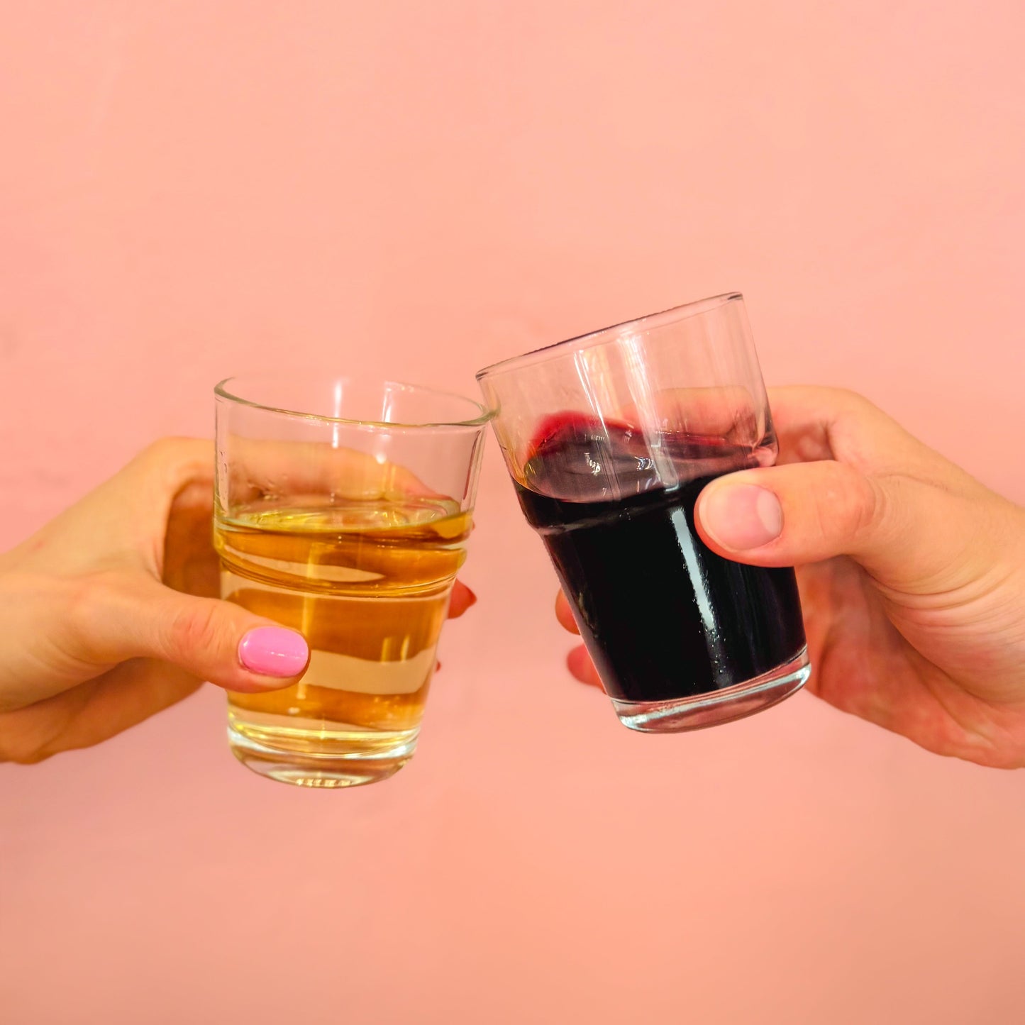 Two hands clinking glasses of red and white wine against a pink background