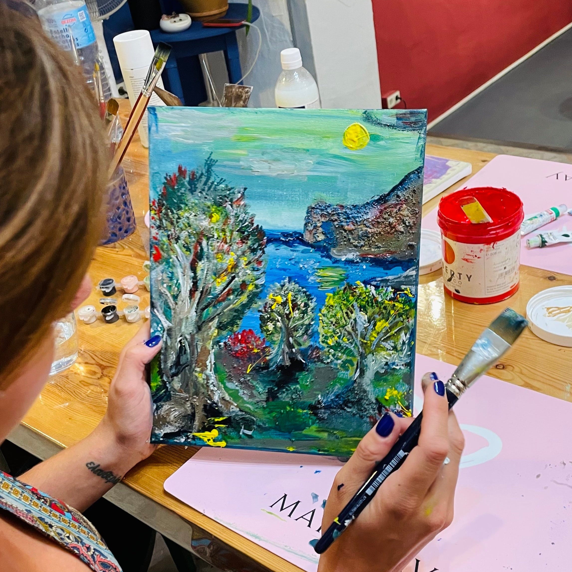 Person holding a colorful painting of a landscape with trees and water, surrounded by art supplies on a table.