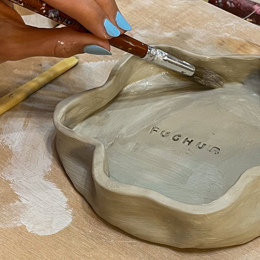 Person working on a ceramic dish with a tool, surrounded by clay and tools on a wooden surface.